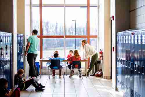 Two teachers stand by a long desk in front of a window where three students sit. The walls in the foreground are lined with blue lockers. Two students sit agains the lockers on the left in the foreground.